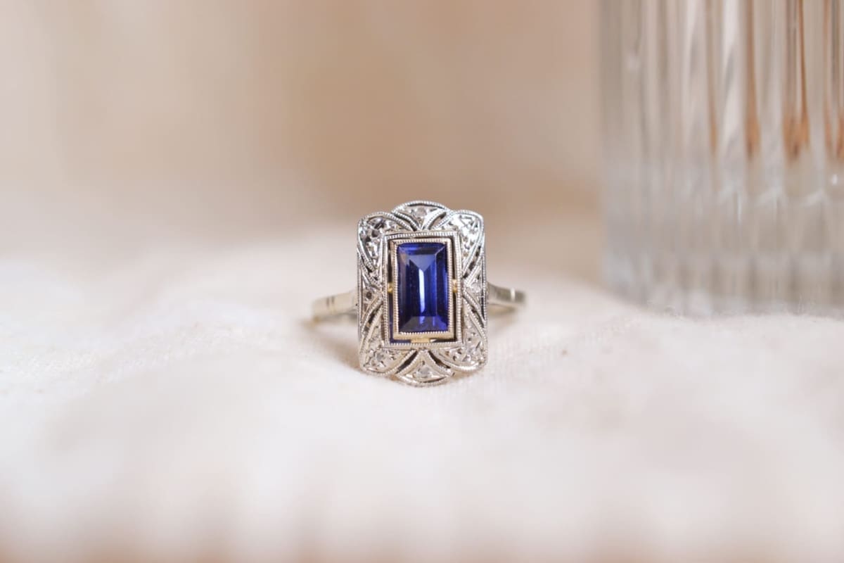 Bague style Art Déco rectangulaire en Or blanc 18 carats et Platine ornée d'un Saphir Verneuil taille à degrés dans un entourage stylisé ponctué de roses de Diamants